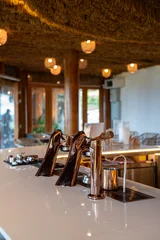 Fototapete Gastronomie Modern tropical cafe interior featuring a pristine white counter, stylish patterned bar stools, and sleek metallic espresso taps under a rustic woven ceiling decorated with warm pendant lights.  © Yellow Boat