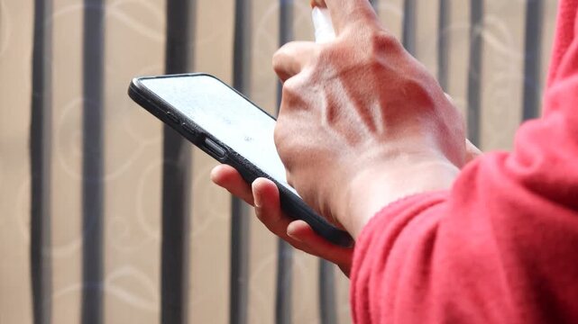Asian man's hand spraying sanitizer and wiping a smartphone screen with a microfiber cloth. Cleaning and disinfecting a mobile phone to remove germs and bacteria.