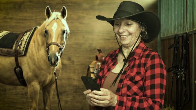 Senior cowgirl using smartphone beside a saddled palomino horse inside a stable. Modern ranch lifestyle blending technology and western tradition. 4K.