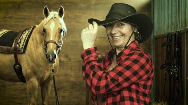 Smiling senior cowgirl tipping her hat beside a saddled palomino horse inside a stable. Western ranch lifestyle and equestrian portrait. 4K.