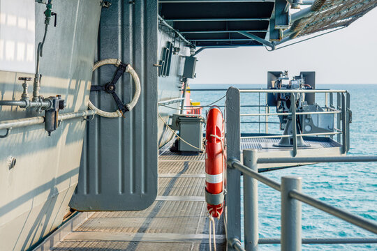 Naval Warship Deck Walkway with Lifebuoy and Anti Aircraft Gun. Outdoor walkway on naval warship deck featuring a red lifebuoy ring, open hatch and anti-aircraft gun station mounted over ocean waters.