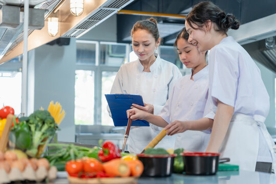 Chef teacher supervising young students cooking in kitchen, Culinary instructor writing on clipboard while kids cooking, Female chef guiding young girls learning how to cook in modern kitchen class