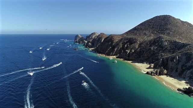Drone flies over the water following the final stretch of land leading toward the Arch of Cabo San Lucas, not yet visible in frame, as boats move across the water below beneath the open sky.
