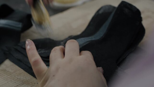 Hands applying adhesive to black shoe uppers with a brush in a workshop setting