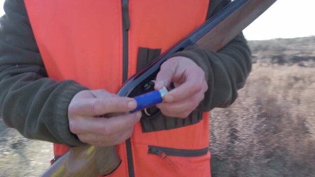 Close-up of hunter wearing orange safety vest holding shotgun cartridge outdoors. Concept for hunting season, shooting sport, ammunition inspection, and field gear.