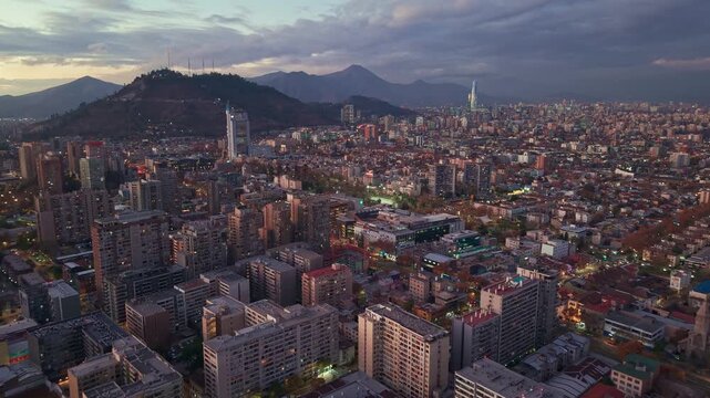 Advancing flight over dense apartment towers and rooftops toward Cerro San Cristobal and distant mountains at dusk. Santiago, Chile