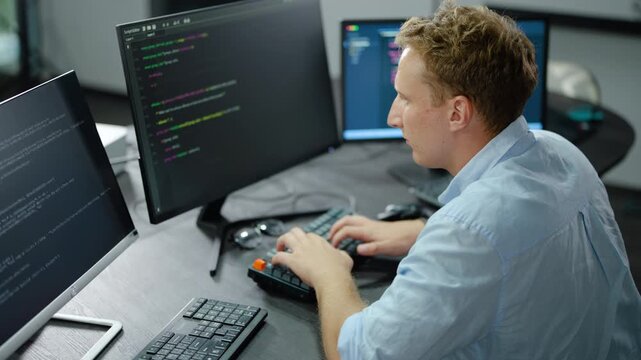 Focused male software developer writing code on multiple computer monitors in a dark modern office. IT professional programmer typing on keyboard working on a cyber security software project.