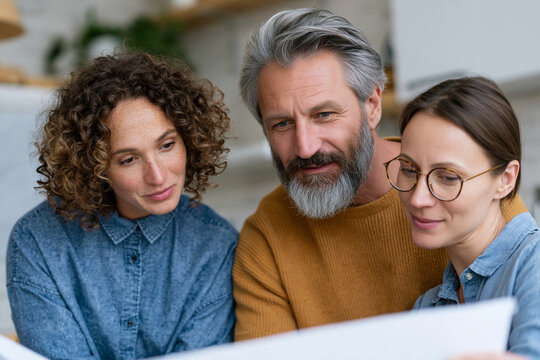 Three people share a quiet moment together. They lean close, focused on a tablet screen. Their expressions show calm interest and mutual trust. Warm lighting enhances the cozy, collaborative mood