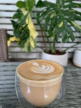 Artful Latte in Bloom: A perfectly crafted latte with intricate foam art sits in a clear glass cup, surrounded by lush green foliage, evoking a serene and artful coffee experience.