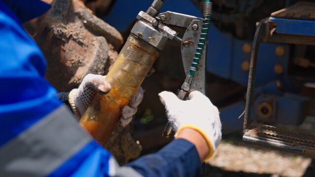 Asian male mechanic engineer in safety helmet using grease gun lubricating heavy tractor machinery parts outdoor. Professional industrial vehicle maintenance and agricultural service.