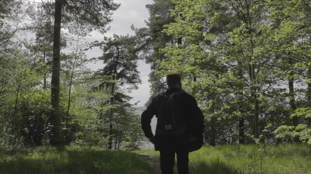 Young male traveler with backpack standing in Bygdoy forest surrounded by spring trees walking along path out to water