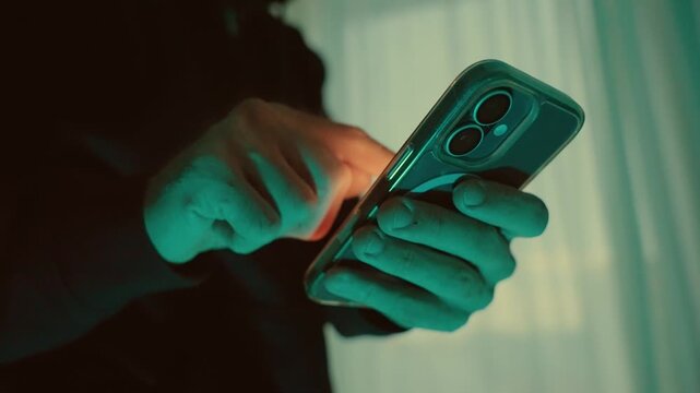 Mysterious male hands holding smartphone and scrolling through social media feed in dark room illuminated by the device's screen. Hacker sending and texting or malware spy app and virus