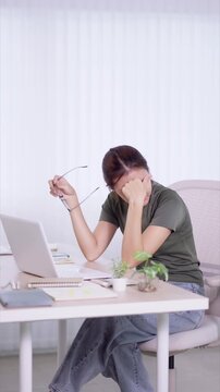 Stressed Asian woman removing glasses while rubbing eyes from fatigue working on laptop at home office desk