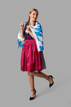 Beautiful happy Octoberfest waitress with Bavarian flag on grey background