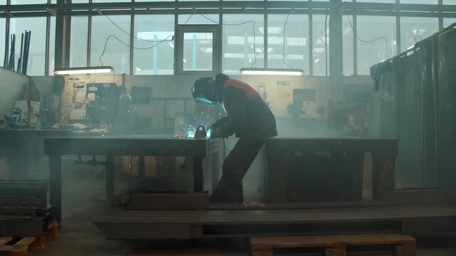 Welder works diligently in an industrial workshop, using specialized tools to fuse metal components together while clouds of smoke fill the interior space of the large manufacturing factory facility.