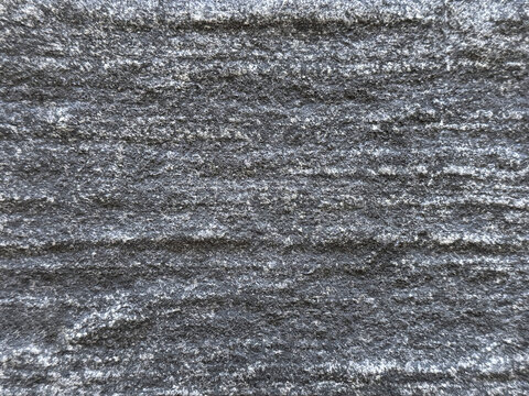 Close up texture of dark grey fluted stone featuring linear horizontal grooves, coarse granular mineral specks, and a rugged chiseled surface.