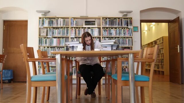 Lonely student studies at a library table surrounded by empty chairs and piles of books showing isolation and pressure during demanding exam preparation without much help