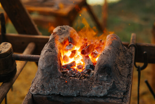 A forge rests under an open sky, filled with glowing coals and flames. Tools and equipment for blacksmithing are placed around, suggesting an active crafting scene