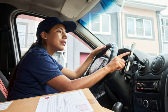 Hispanic young adult female courier sitting in delivery van and using smartphone GPS navigation. Image represents logistics and e-commerce shipping services, low angle shot