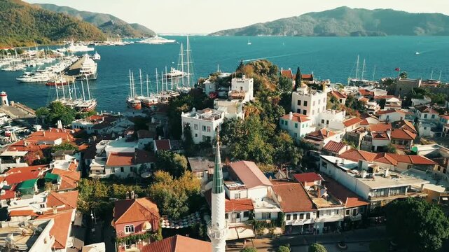 Aerial drone shot overlooking Symi Town with a white minaret rising among red-roofed buildings as sailboats fill the harbor surrounded by rugged hills in Greece