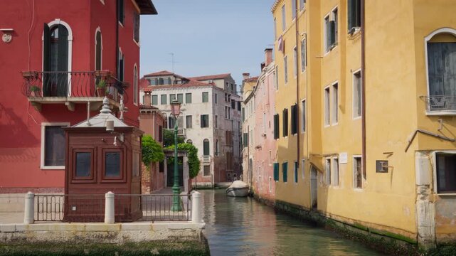 A narrow canal runs between colorful historic buildings, with a small moored boat, waterfront kiosk and lamppost in a residential area of Venice, Italy.