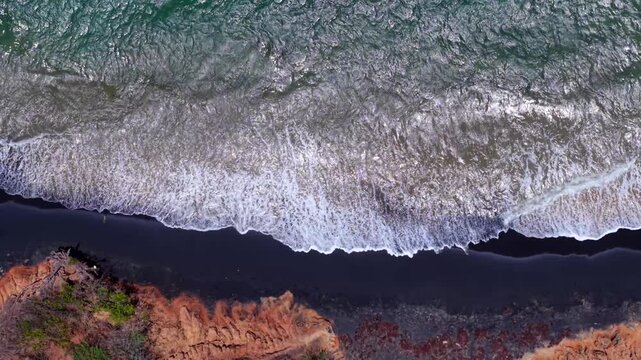 Top-down approaching drone view over foamy Caribbean waves roll onto the Black Sand Beach below rugged red cliffs, Vieques, Puerto Rico