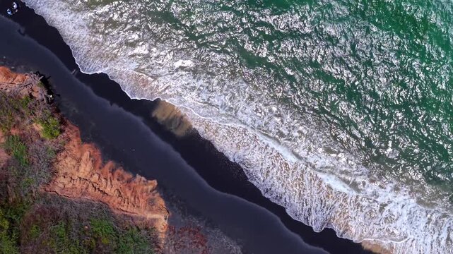 Dynamic spiral drone fly over Black Sand Beach shoreline with foamy Caribbean Sea surf and reddish volcanic coastal cliffs, Vieques, Puerto Rico