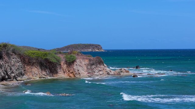 Backward drone fly of rocky cliffs meet turquoise Caribbean waves along the Vieques shoreline under a clear blue sky in summer, Puerto Rico