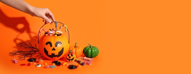 Fototapeta premium Hand reaching for halloween candy in a jack-o'-lantern bucket with pumpkins and sweets on an orange background, Pumpkin, Holiday