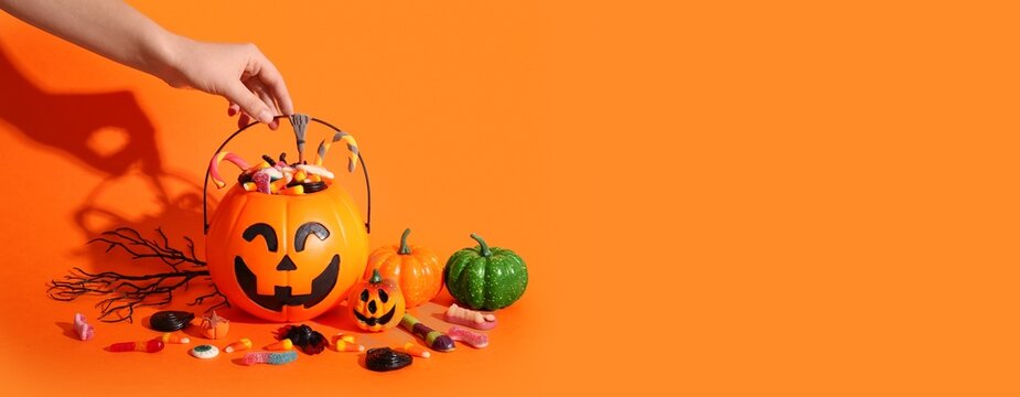 Hand reaching for halloween candy in a jack-o'-lantern bucket with pumpkins and sweets on an orange background, Pumpkin, Holiday