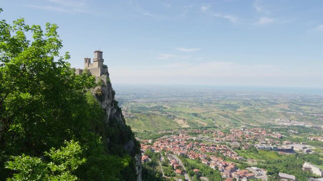A medieval stone fortress crowns a steep cliff above villages, patchwork fields and rolling countryside in San Marino under a bright blue sky.