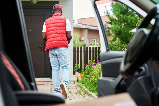 African American young adult courier walking toward house door to deliver package, seen from inside car. Image represents e-commerce shipping and home delivery services