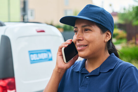 Hispanic young adult female courier talking on smartphone near delivery van. Image represents logistics, customer service, and mobile business communication, close up shot