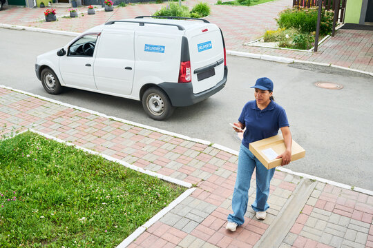 Hispanic female courier holding smartphone and cardboard box walking near delivery van. High angle shot representing e-commerce logistics and shipping services for commercial marketing