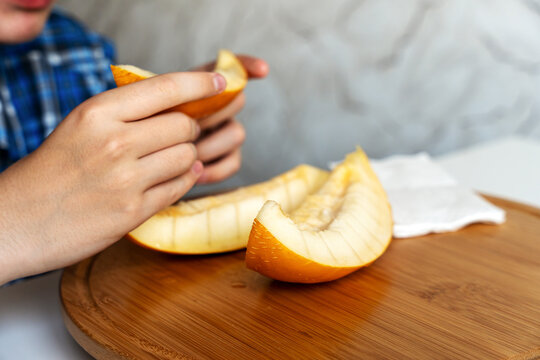 A close-up of a teenage boy in a blue plaid shirt sitting at a table and eating fresh melon. He is holding a juicy slice of yellow melon, which is neatly cut into bite-sized pieces on a cutting board.
