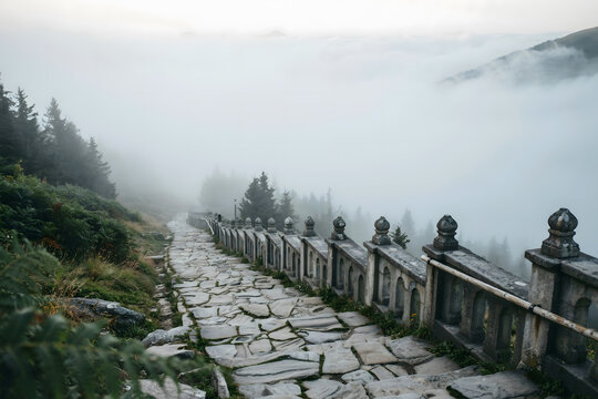 A stone hiking trail with a decorative stone railing winding through a foggy mountain forest landscape during an early morning atmosphere