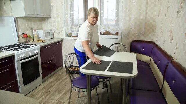 Elderly Caucasian woman preparing dining table in bright domestic kitchen interior with modern appliances and purple furniture, placing dark placemats on white surface