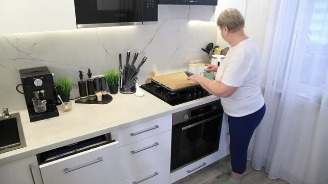 Older Caucasian woman preparing baking tray with parchment paper on a gas hob in a sleek modern kitchen interior, ready for cooking process, domestic scene of food preparation activity