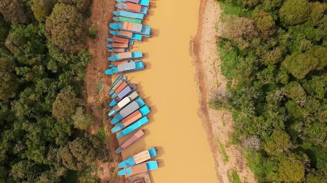 Cinematic 4K aerial drone footage of Tonle Sap floating village in Cambodia, showcasing a winding muddy canal lined with traditional stilt houses, colorful fishing boats, lush green countryside