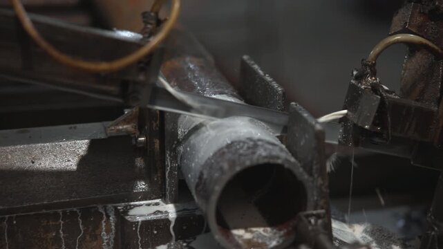 Close view of bandsaw cutting cylindrical steel pipe with coolant spraying across rusted machine bed inside industrial workshop, showing precise metalworking, wet surfaces, and heavy fabrication