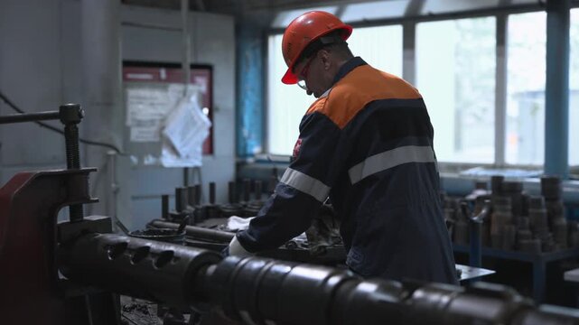 Industrial mechanic in orange hardhat and safety glasses inspecting oily metal component at factory workshop bench, wearing gloves and holding rag beside vise under bright daylight