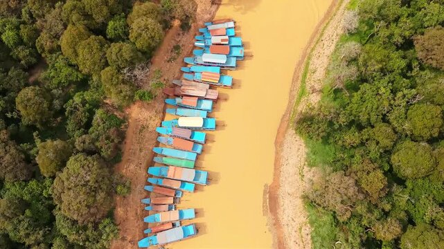 Cinematic 4K aerial drone footage of Tonle Sap floating village in Cambodia, showcasing a winding muddy canal lined with traditional stilt houses, colorful fishing boats, lush green countryside