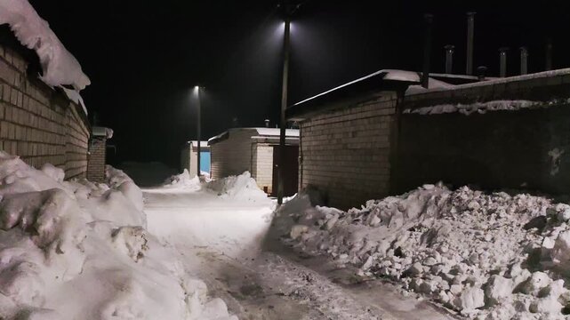 A snow-covered alleyway in a garage co-op at night, illuminated by streetlights, casts long shadows across the snowdrifts and brick garages, creating a cold, deserted winter scene.