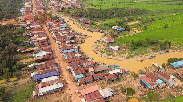 Cinematic 4K aerial drone footage of Tonle Sap floating village in Cambodia, showcasing a winding muddy canal lined with traditional stilt houses, colorful fishing boats, lush green countryside