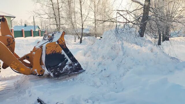 Yellow excavator bucket removing large snowdrift from winter road, heavy equipment clearing white snow pile near residential houses and bare trees during cold frosty day