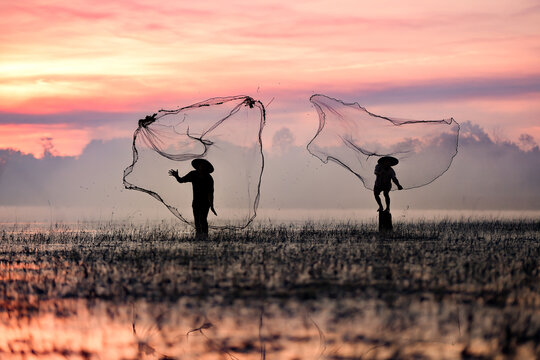 Silhouette of traditional fishermen casting net at golden sunrise in Sakon Nakhon, Isan, Thailand. Capturing authentic rural Asian lifestyle, culture, and fishing for daily sustenance in tranquil lake