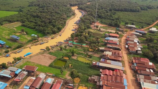Cinematic 4K aerial drone footage of Tonle Sap floating village in Cambodia, showcasing a winding muddy canal lined with traditional stilt houses, colorful fishing boats, lush green countryside