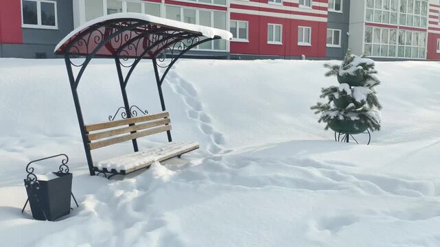 Winter landscape featuring snow-covered park bench with metal canopy, small evergreen tree, trash can, and footprints in deep snow with residential buildings background under bright sunlight