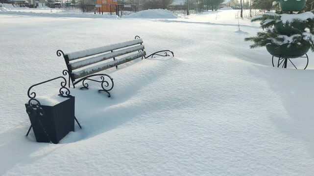 Empty park bench and trash bin covered in deep pristine white snow on a clear winter day, long shadows cast by bright sun across a quiet outdoor landscape