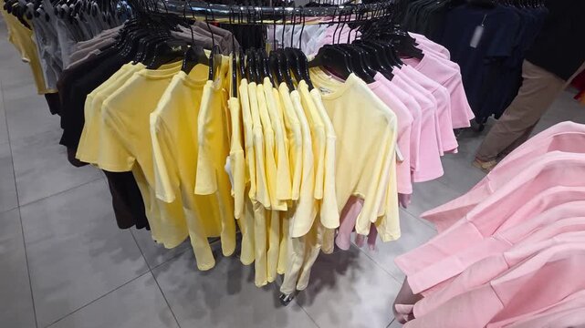 Circular clothing rack display showing rows of yellow and pink t-shirts for sale in a retail store, hand of a shopper browsing apparel